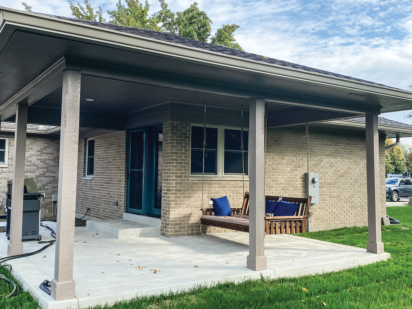 A covered backyard patio with concrete flooring, a porch swing, a grill, and outdoor seating, attached to a brick house.