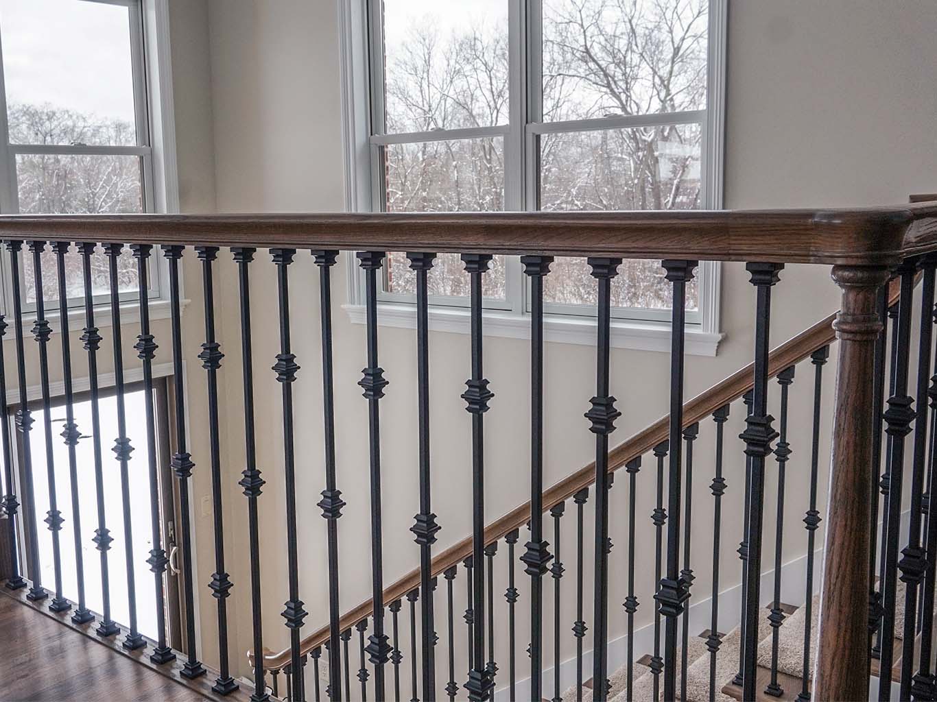 Wooden staircase railing with black metal balusters, positioned next to large windows showing a snowy outdoor landscape.