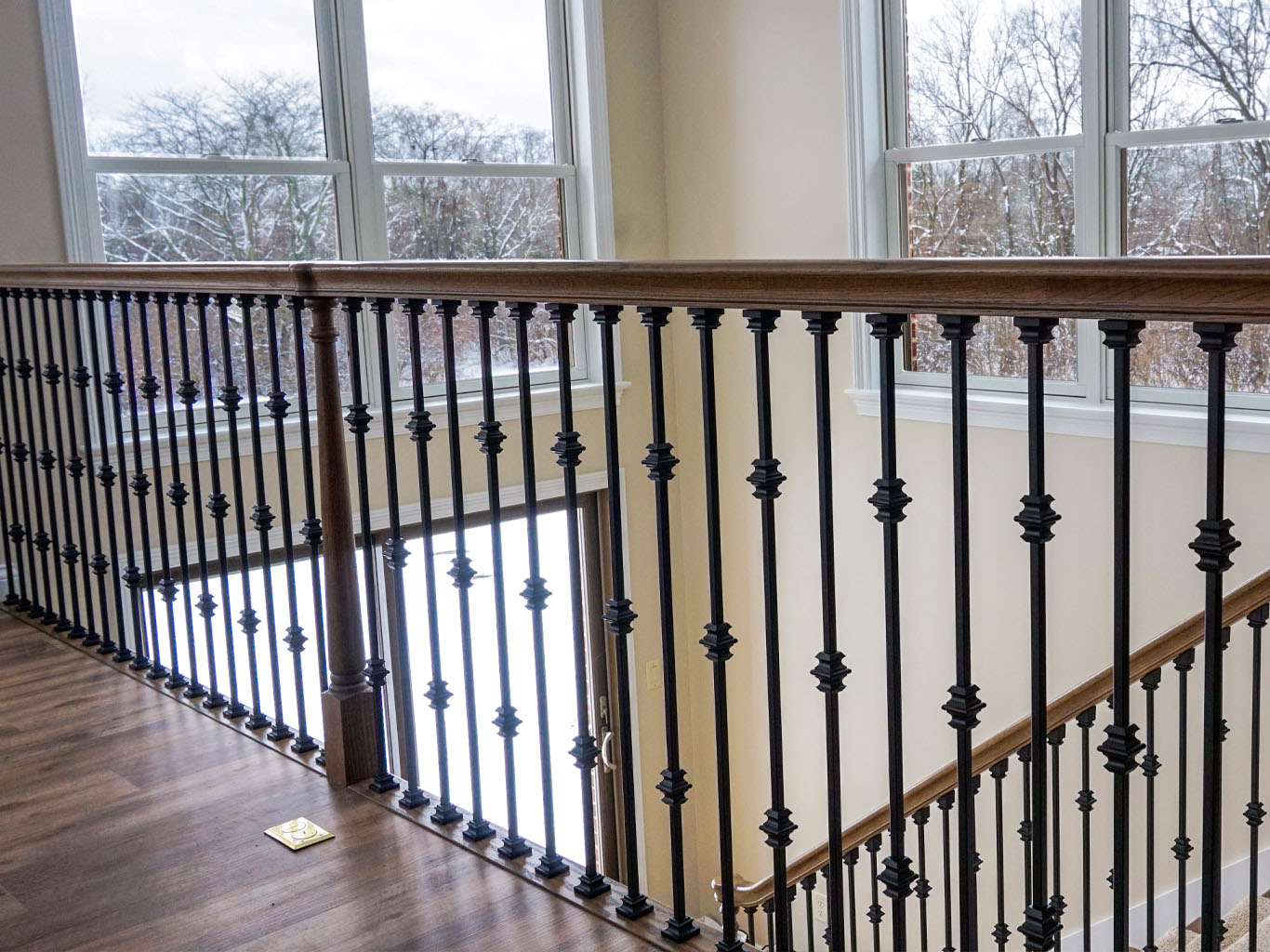 A wooden handrail with black metal balusters lines a staircase landing, adjacent to large windows showing a snowy outdoor scene.