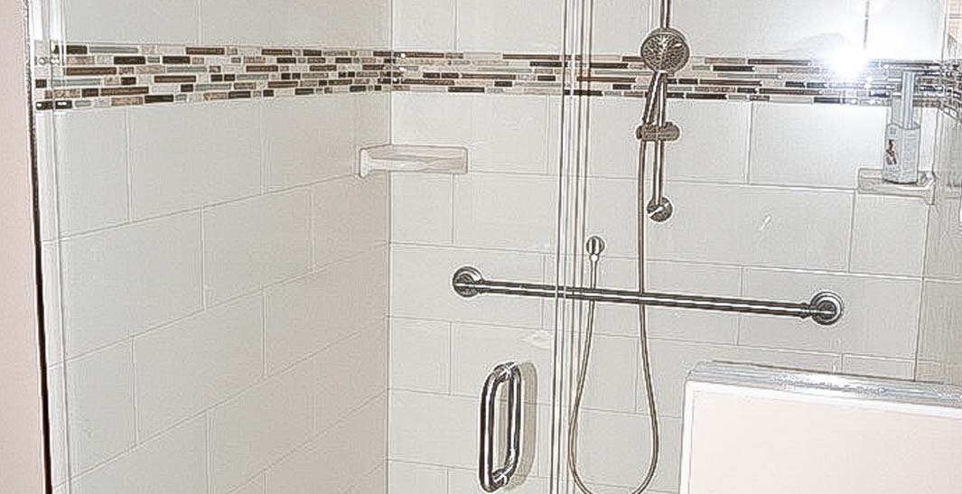 A modern shower with white tiles, a decorative border, a glass door, a metal grab bar, and a handheld showerhead.