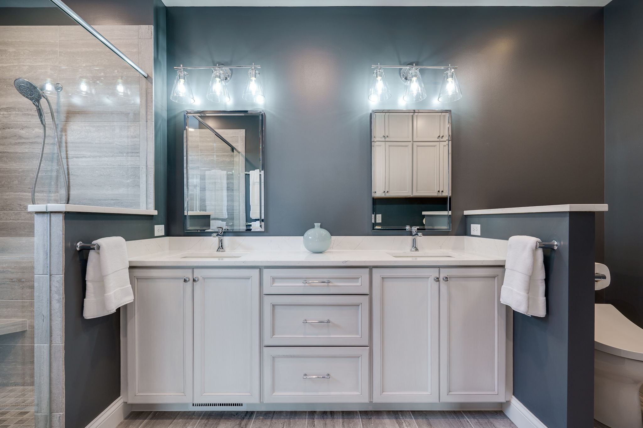 Modern bathroom remodel in Dayton, Ohio, featuring a double sink vanity, two mirrors, overhead lighting, a glass-enclosed shower on the left, and a partially visible toilet on the right.