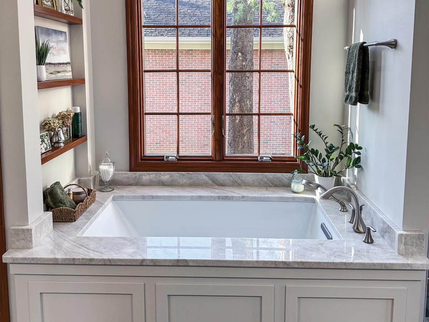 A white soaking tub with a marble surround is positioned beneath a large window; shelves with decor and towels are on the left, and a plant sits by the faucet.