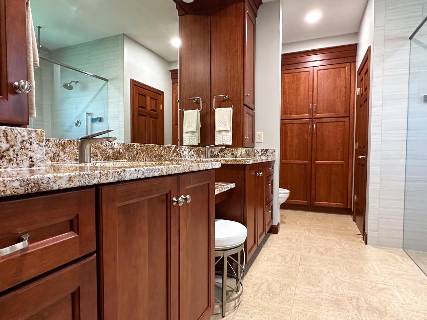 Bathroom with wood cabinets, granite countertop, dual sinks, mirrors, a walk-in shower with glass door, and beige tile flooring. Towels hang between the sinks.
