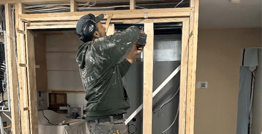 A person wearing a cap and hoodie is using a power drill on a wooden frame in a construction area.