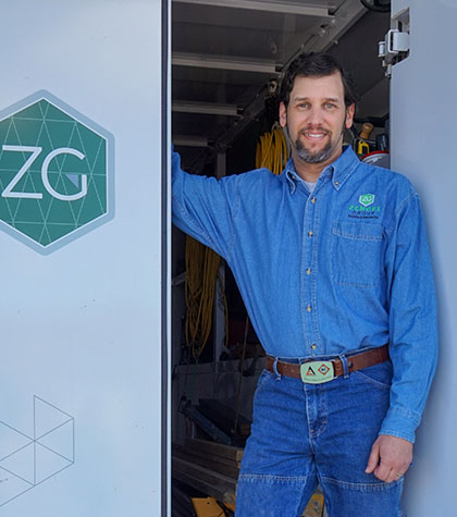 A person in a blue denim shirt stands at the open door of a vehicle with "ZG" logo, smiling with one hand resting on the door.