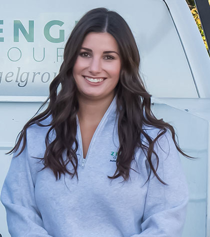 A woman with long, dark hair smiles, wearing a light gray pullover, standing in front of a vehicle with partial text visible.