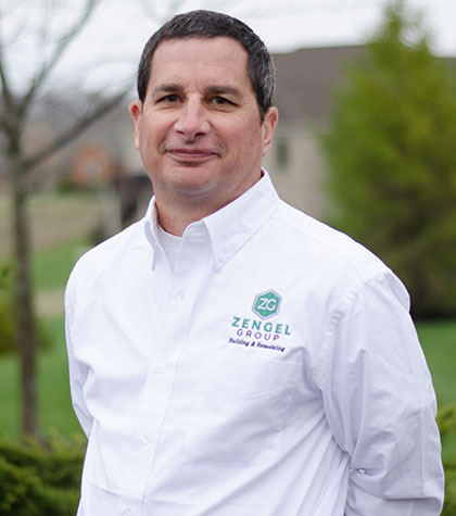 Man in a white shirt with "Zengel Group" logo stands outdoors with blurred greenery and buildings in the background.