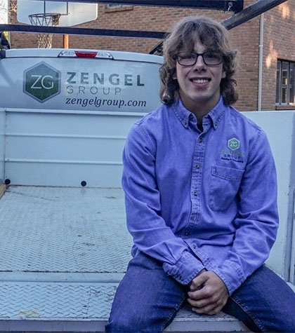 Person seated on a truck bed wearing a purple Zengel Group shirt. Brick building and basketball hoop in the background.