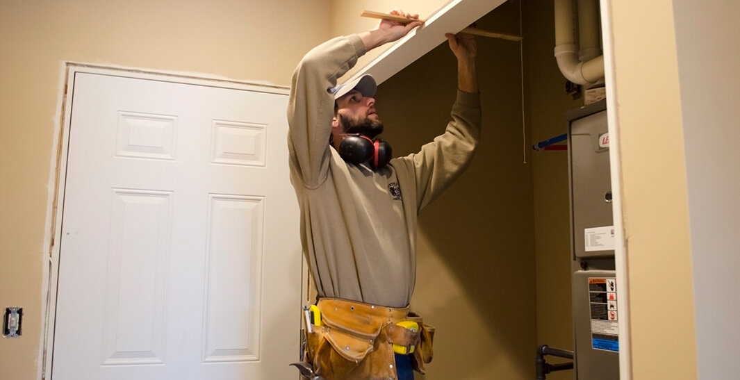 A person in a work uniform and tool belt installs a panel near a furnace in a utility room.