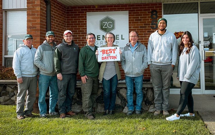 A group of eight people standing in front of a brick building with a sign reading "Best of Dayton." They are smiling and casually dressed.