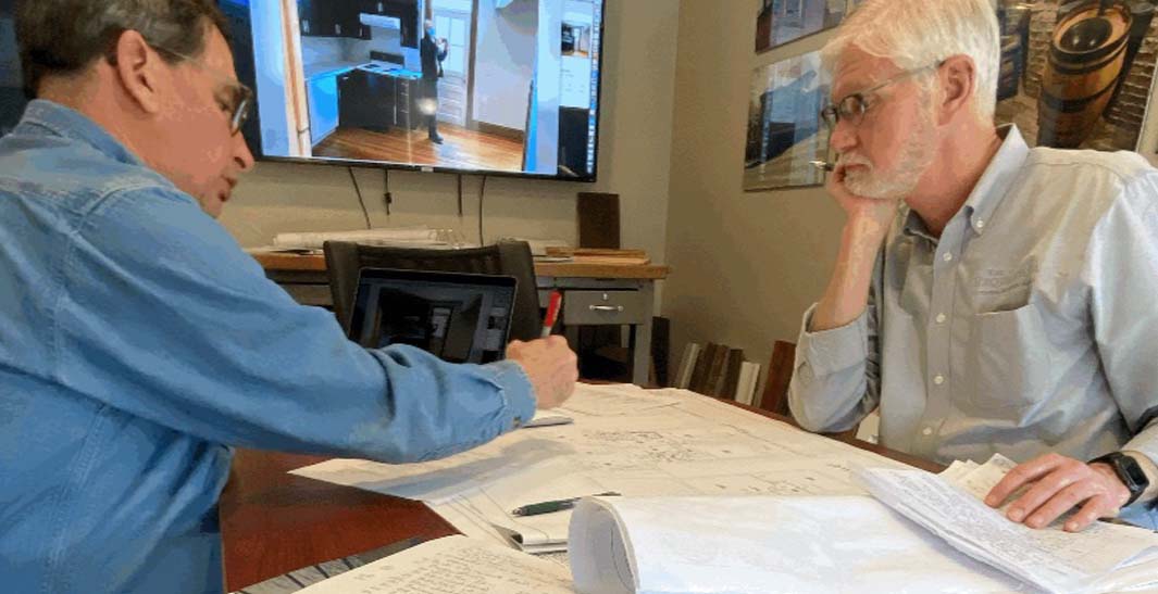 Two men are discussing blueprints at a table. A television in the background displays architectural images.