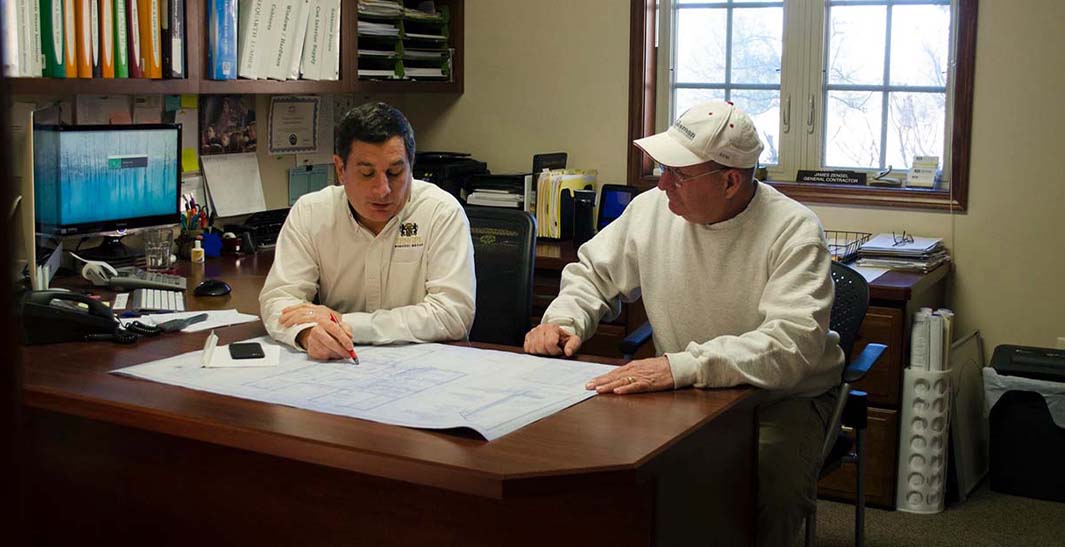 Two men sitting at a desk reviewing architectural plans in an office setting.