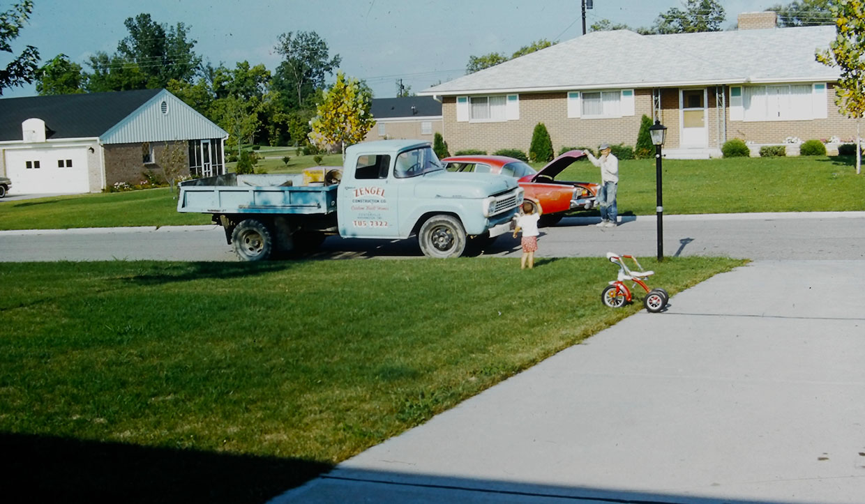 A vintage truck and a car with an open hood are parked on a suburban street. A toddler stands on the sidewalk next to a tricycle. A row of houses and trees are in the background.