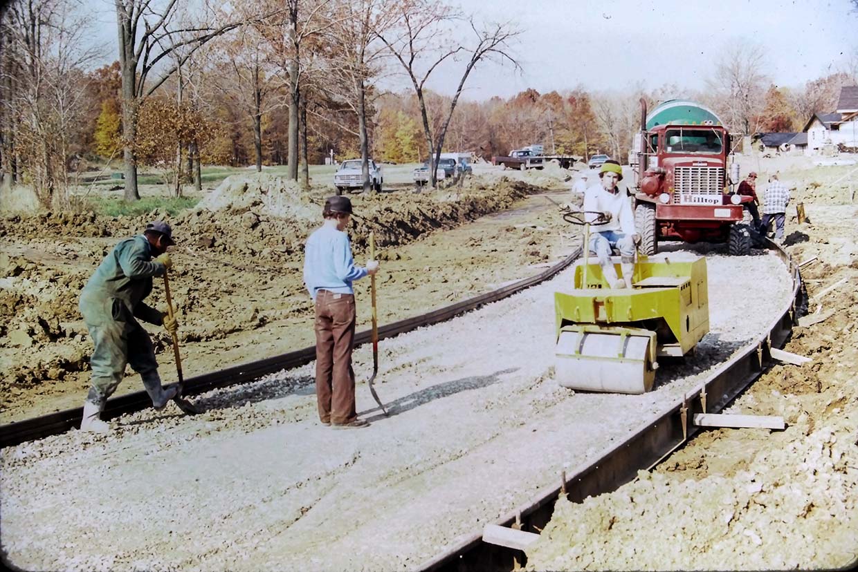 Workers paving a road with gravel using a roller compactor and shovels. A red truck is parked nearby. Trees and construction materials are in the background.