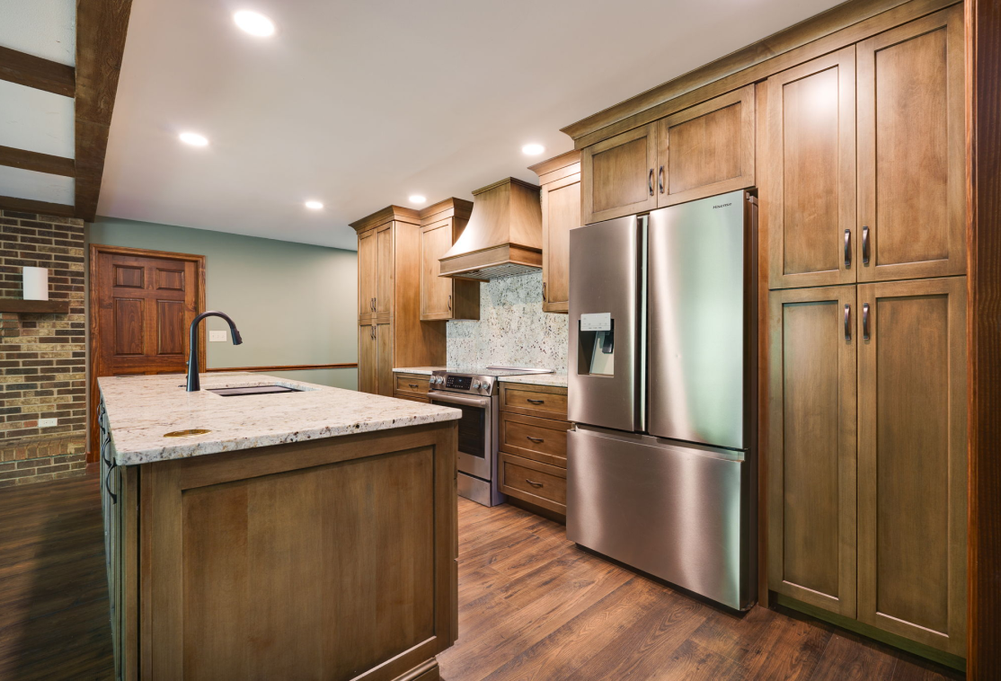 Modern kitchen with wooden cabinets, a stainless steel refrigerator, an island with a sink, and a stove with a range hood. Recessed lighting and hardwood floors complete the room.