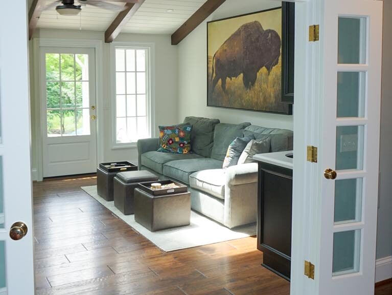 A cozy living room with a sectional sofa, colorful pillow, artwork of a bison, and wooden ceiling beams. The room has large windows and a door leading outside.