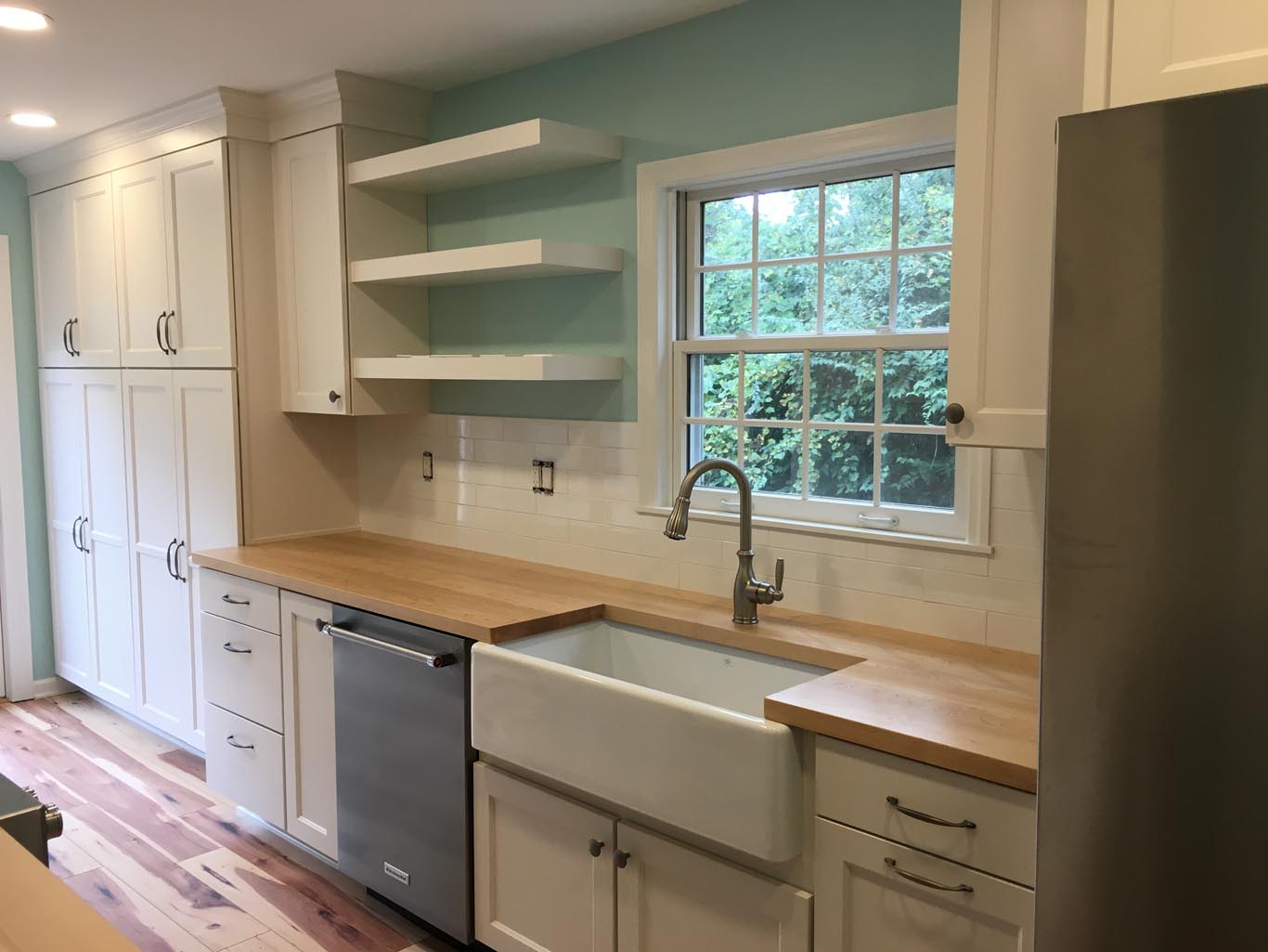 Modern kitchen with light wood countertops, white cabinets, a large farmhouse sink, stainless steel dishwasher, open shelves, and a window overlooking greenery.