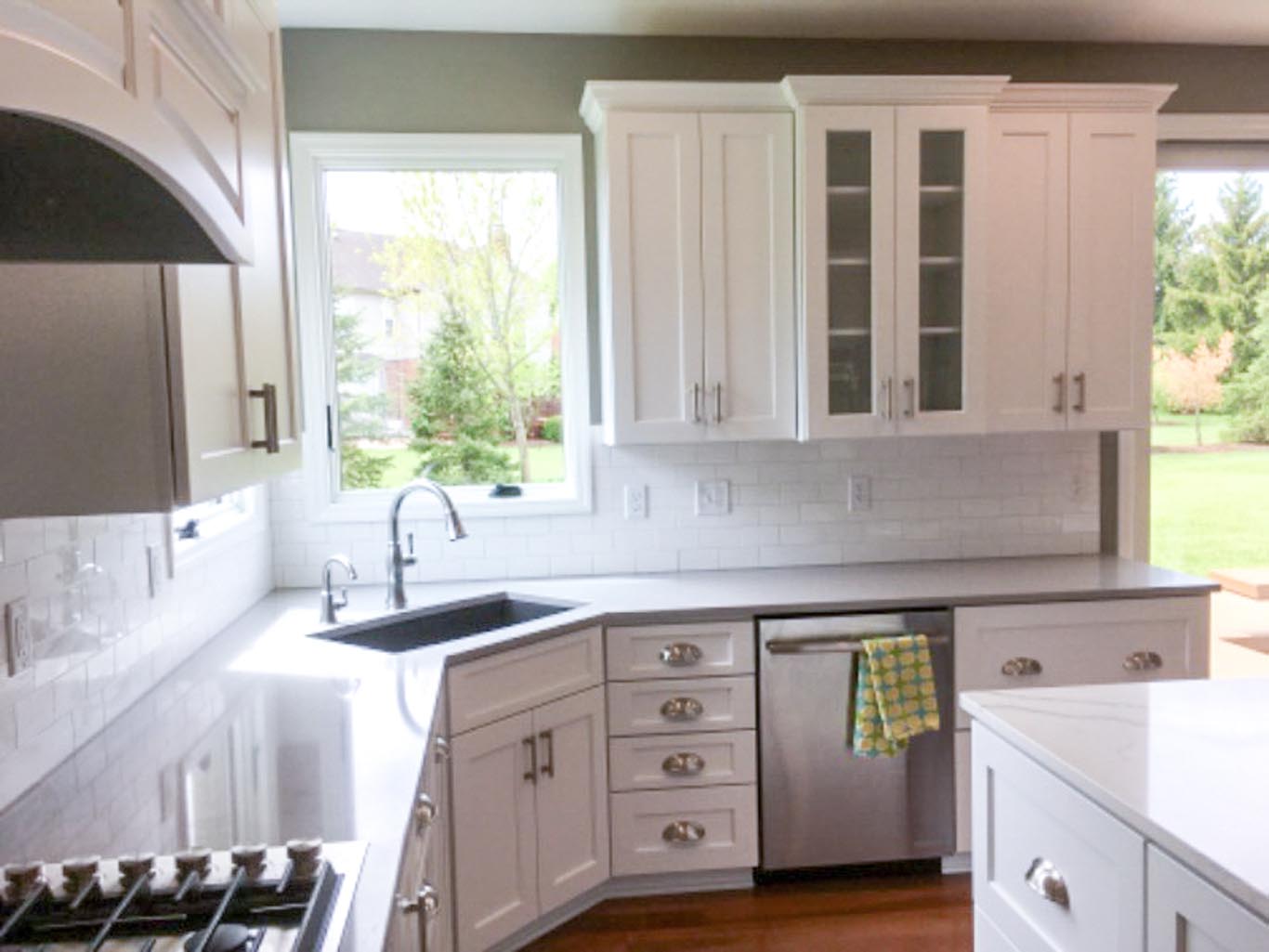 Modern kitchen with white cabinets, stainless steel appliances, a window overlooking a garden, gray countertop, and a sink with faucet. A green checked towel hangs on the dishwasher handle.