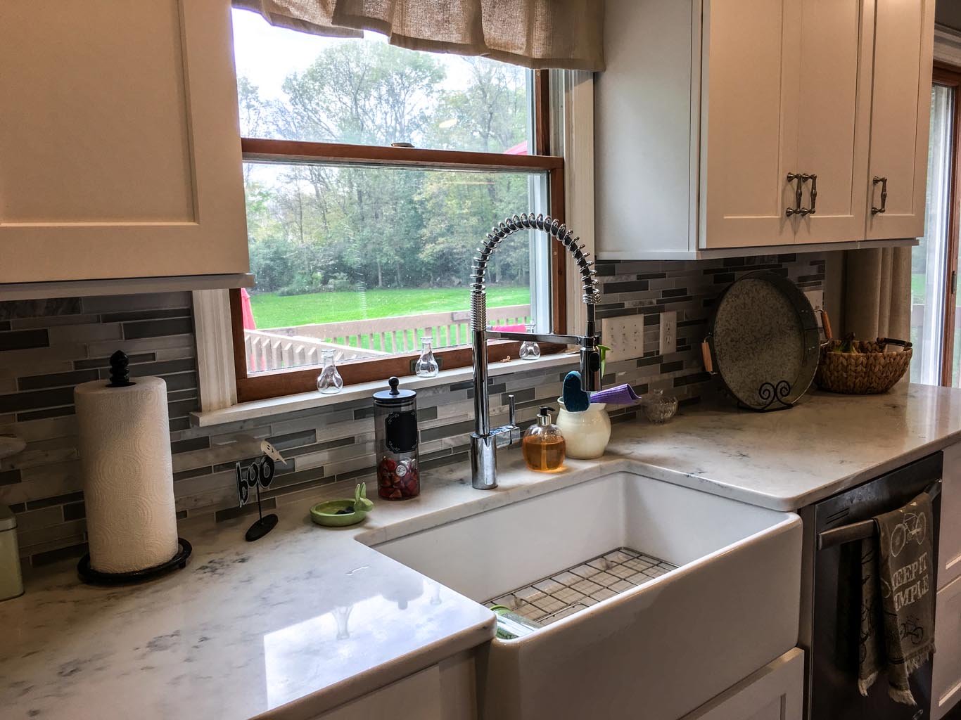 A kitchen interior with a large farmhouse sink, marble countertops, a chrome faucet, and a window overlooking a green outdoor scene. White cabinets and a backsplash decorate the space.