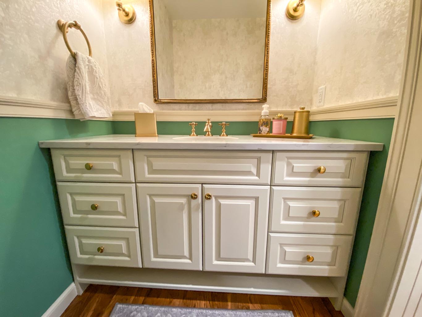 A small bathroom with a white vanity, gold fixtures, a large mirror, and green walls. There is a towel holder, tissue box, and decorative items on the counter.