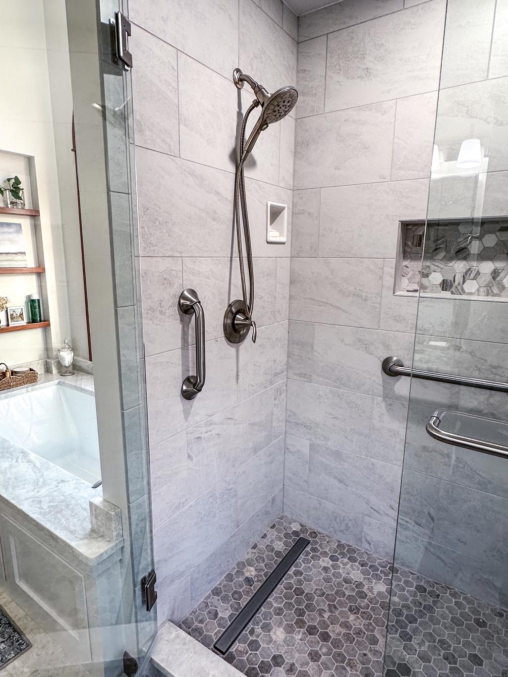 Modern bathroom with a glass-enclosed shower featuring gray tiles, a dual showerhead, and a built-in niche. Adjacent is a bathtub, with shelves and a window in the background.