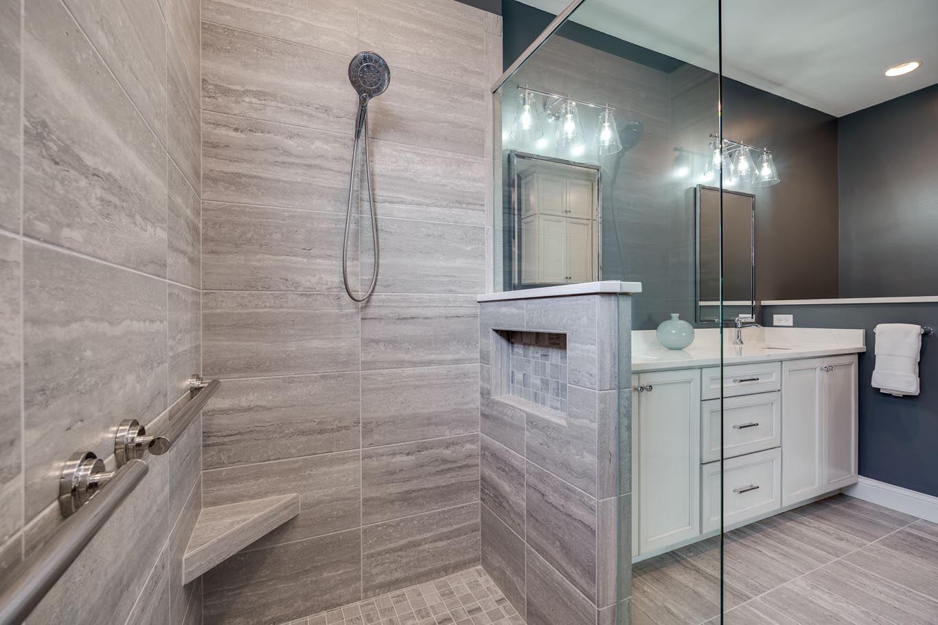 Modern bathroom with a glass-enclosed shower, gray tile walls, built-in bench, and a chrome showerhead. A vanity with a mirror and lighting is visible in the background.