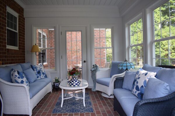Enclosed sunroom with two blue-cushioned wicker sofas, a round table with a checkered vase, brick walls, and large windows showing green foliage outside.