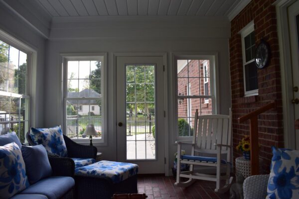 Sunlit porch with blue floral cushions on wicker furniture, a white rocking chair, and brick walls. Large windows and a glass door provide a view of the garden outside.