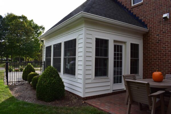 A white sunroom with large windows extends from a red brick house. A wooden patio set with a pumpkin on the table is in the foreground, and a wrought iron fence borders a garden area.