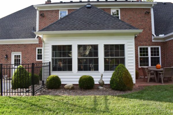 A brick house with a white sunroom featuring three large windows. There is a patio with a table, chairs, and a pumpkin. Shrubs and a metallic fence are in the foreground.
