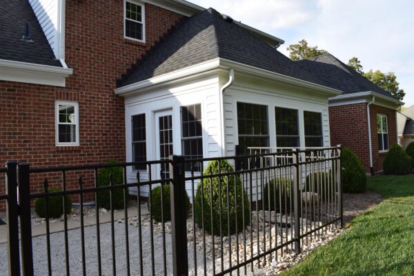 Brick house with a white sunroom, black roof, and black metal fence. Small bushes and gravel surround the area.