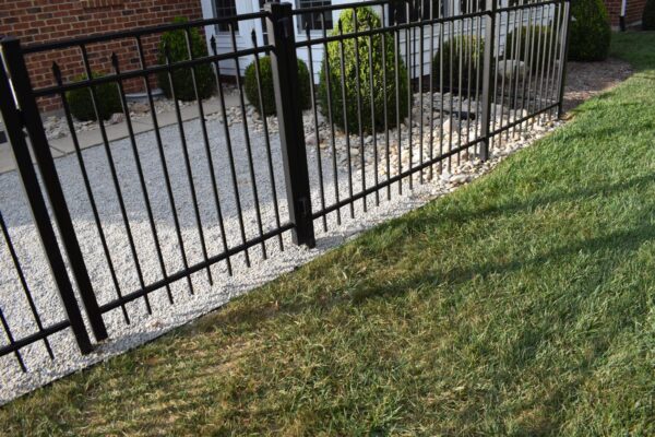 A black metal fence separates a gravel path from a grassy area in front of a brick building with shrubs.