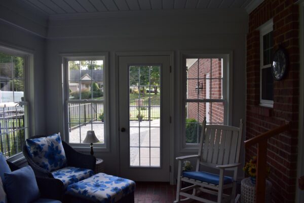 A sunroom with wicker chairs, floral cushions, and a rocking chair. Large windows and a glass door overlook a patio and garden. Brick walls and a clock are visible.