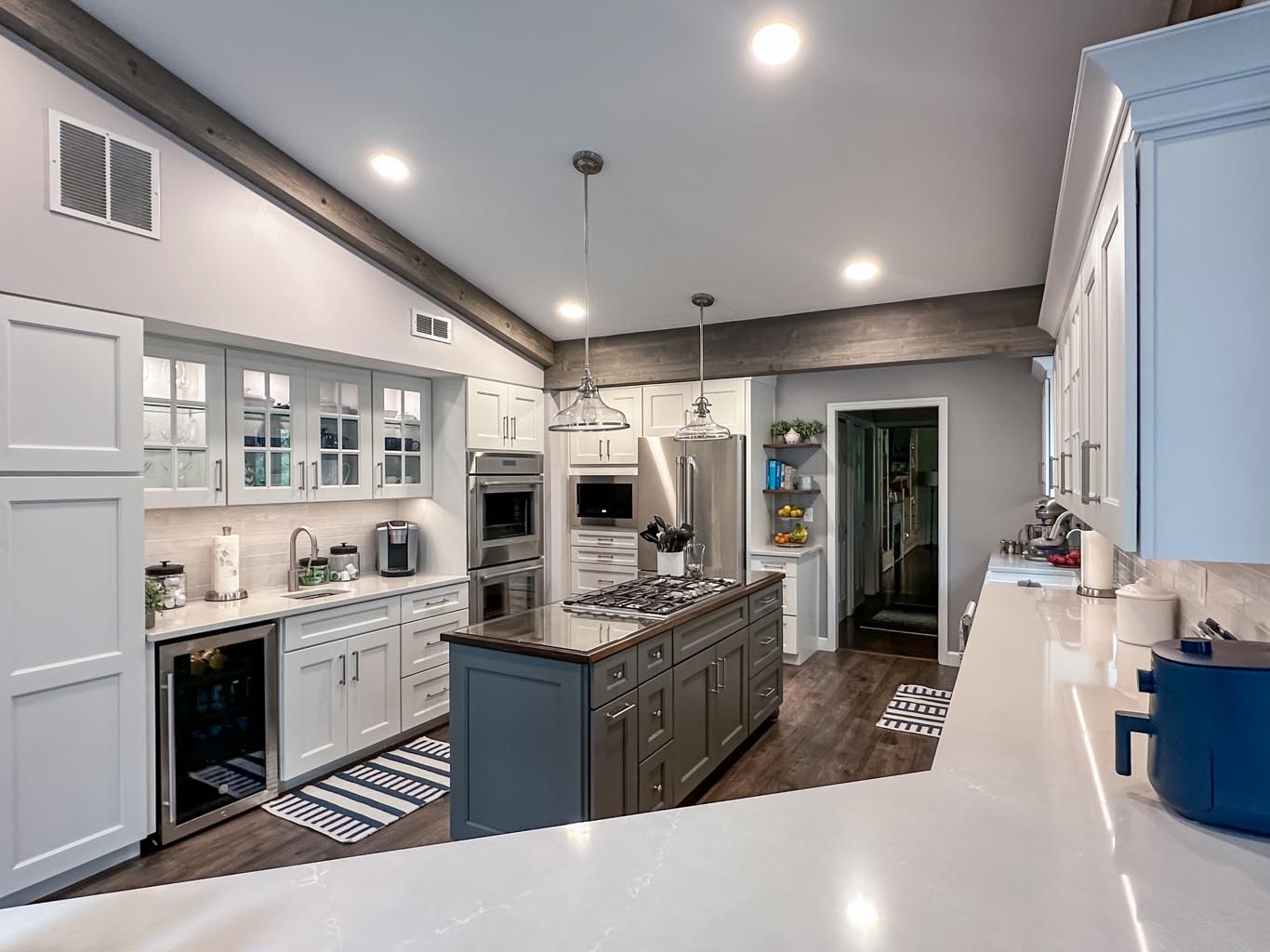 Modern kitchen with white cabinets, an island with a dark finish, stainless steel appliances, and overhead pendant lights. The ceiling features exposed wooden beams.