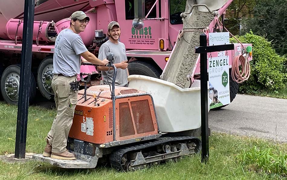 Two workers operate construction equipment, pouring concrete from a cement truck into a small excavator bucket on a grassy area.