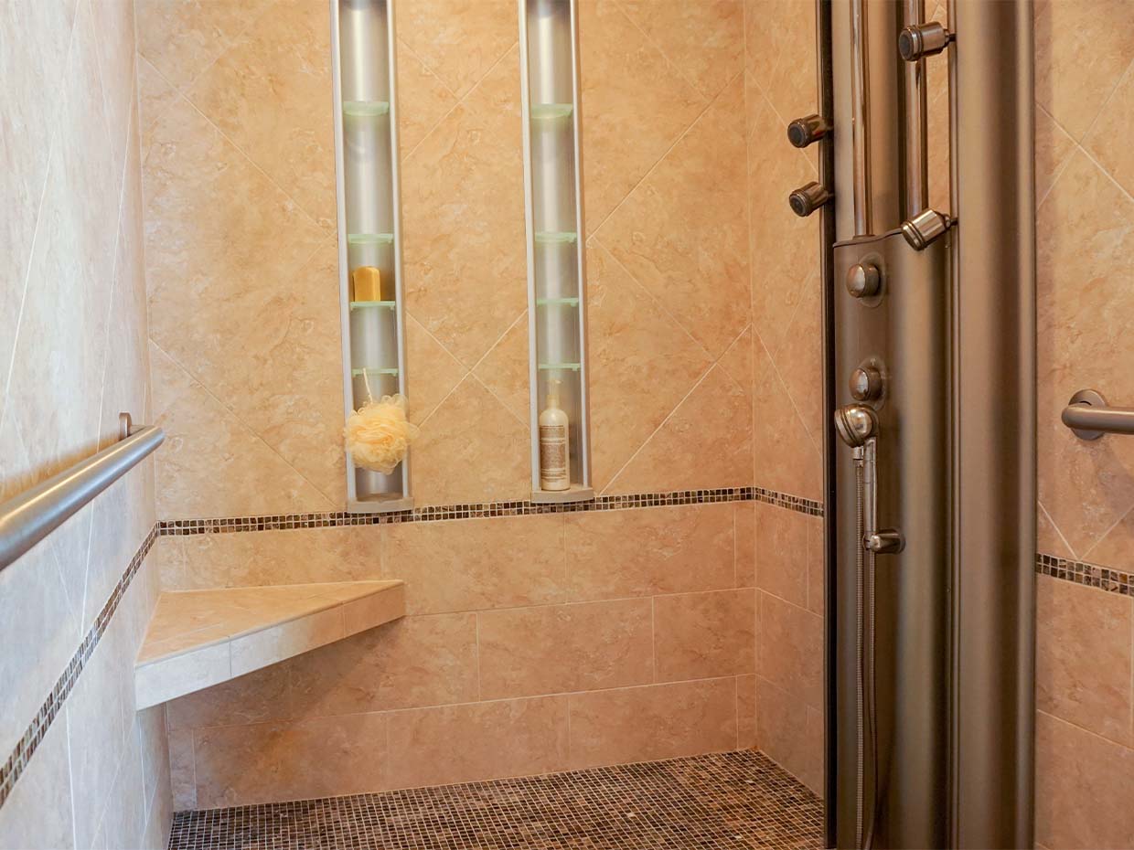 A walk-in shower with beige tiles, a corner seat, and a vertical shower panel featuring multiple showerheads. Built-in shelves hold toiletries, and a grab bar is installed on the wall.