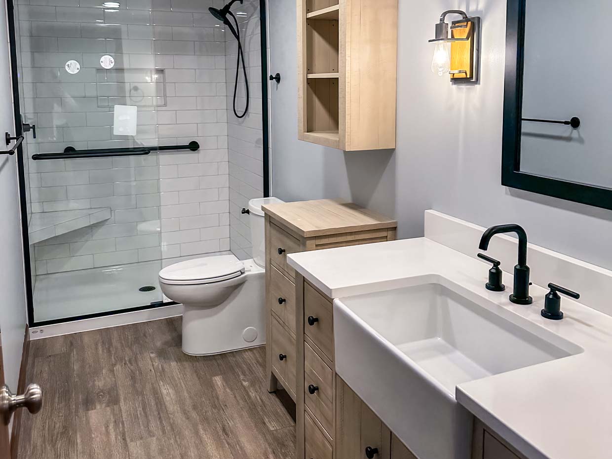 Modern bathroom with a glass-enclosed shower, white toilet, wooden cabinetry, and a sink with black fixtures on a wood floor.