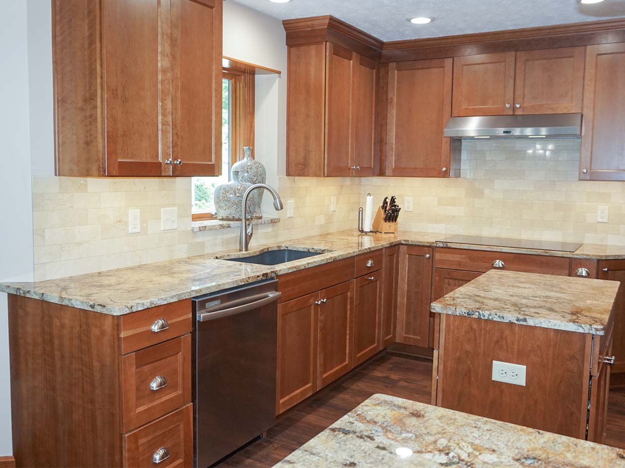 Modern kitchen with wooden cabinets, granite countertops, stainless steel dishwasher, and range hood. Tile backsplash and dark wood flooring complete the design.