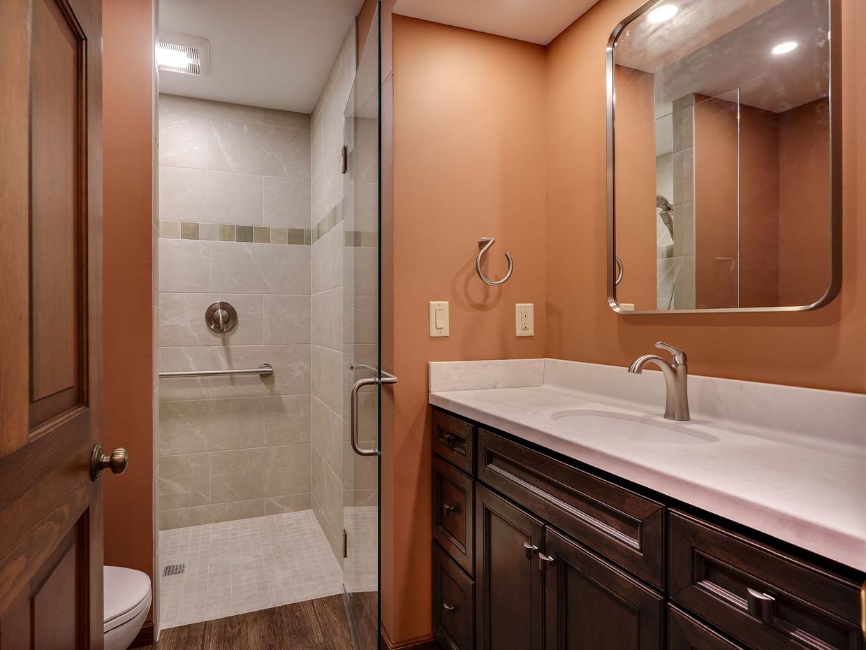 Bathroom with a glass-enclosed shower, wooden cabinets, white countertop, and a large mirror above the sink. Walls are painted in a warm brown tone.