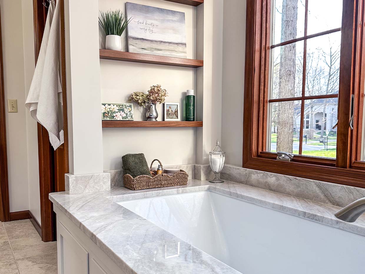 Bathroom with a built-in marble tub, wooden shelves displaying decor, and a large window with a view of trees and houses outside. A towel hangs on the wall.