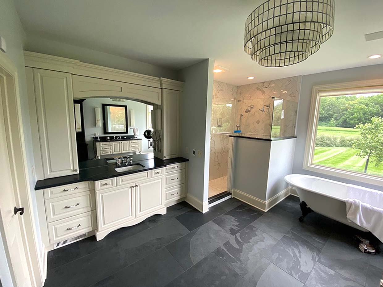 Spacious bathroom with black tiled floor, large vanity, glass-enclosed shower, freestanding bathtub, and a view of a grassy landscape through a window.