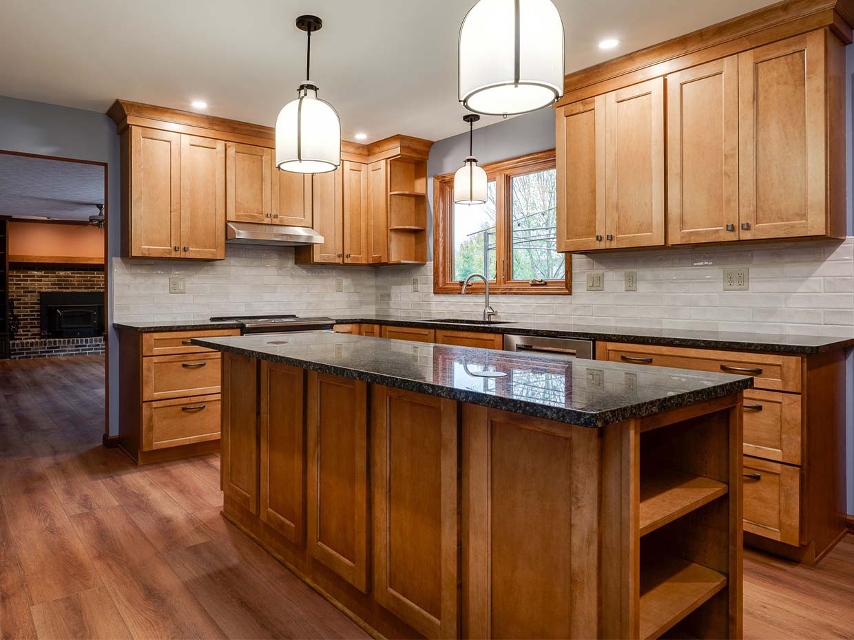 A modern kitchen with wooden cabinets, a large island with a black countertop, pendant lights, and a view into the living area with a brick fireplace.