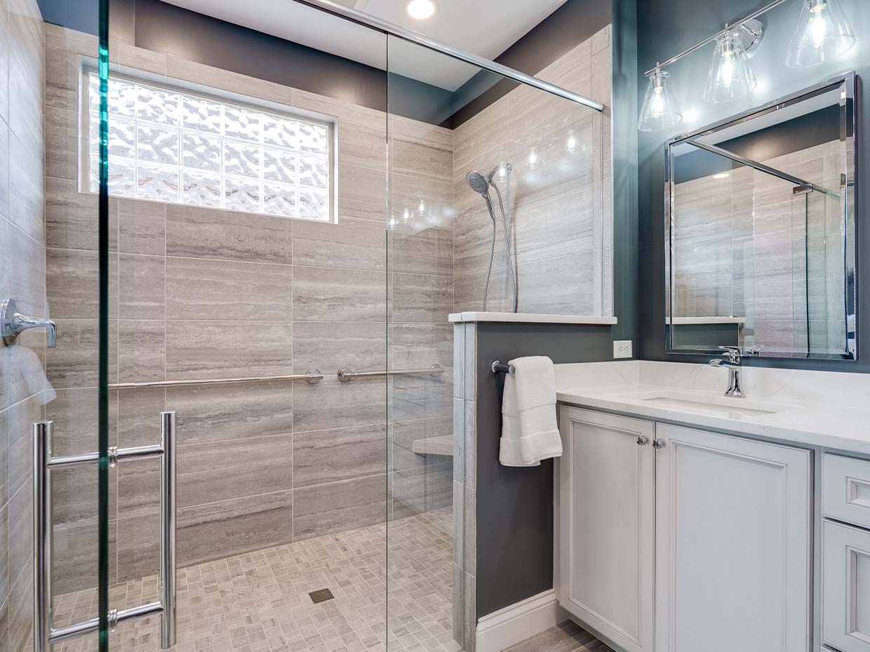 Modern bathroom with a glass shower enclosure, gray tile walls, and a white vanity. Three light fixtures are above the mirror, and a towel hangs on a bar near the shower.