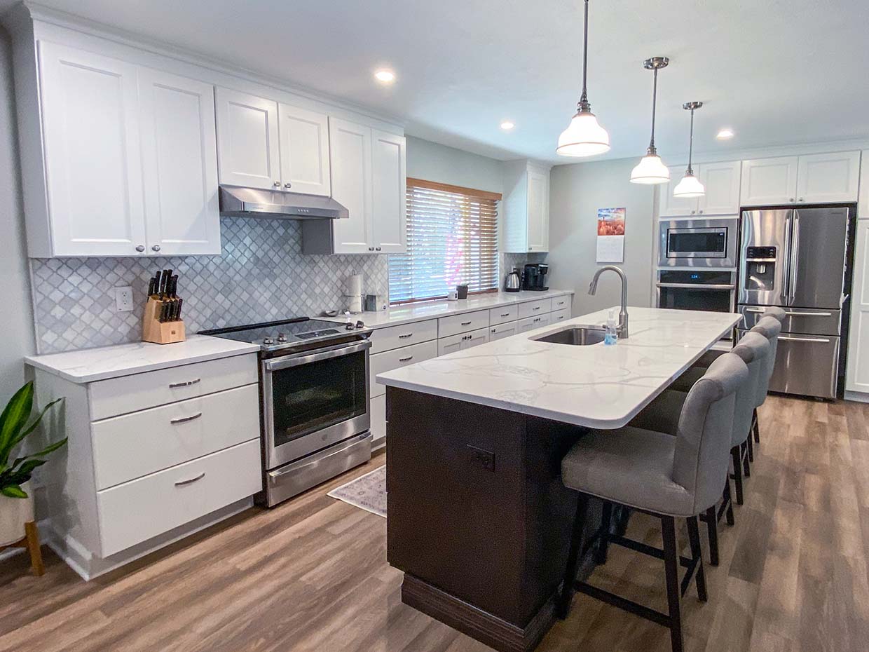 A modern kitchen with white cabinets, marble countertops, pendant lights, stainless steel appliances, and a dark island with gray barstools.