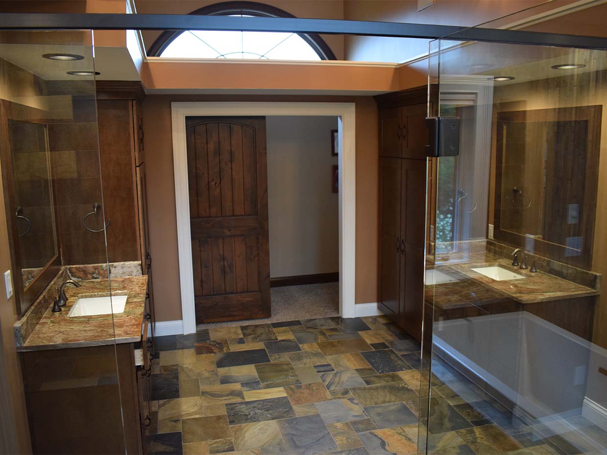 Spacious bathroom with a dual-sink vanity, wooden cabinetry, a mirror, a glass shower enclosure, slate tile flooring, and a wooden door. An arched window above provides natural light.