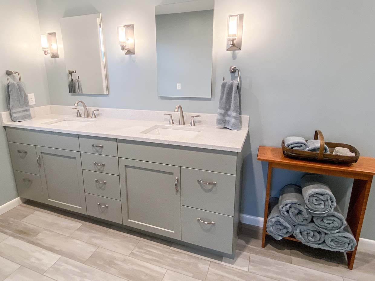 A bathroom with a double sink vanity, two mirrors, wall sconces, and a wooden shelf holding rolled towels.