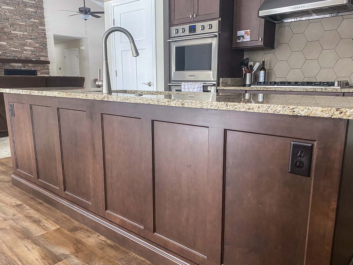 Kitchen island with a granite countertop, stainless steel faucet, and dark wood paneling. Oven and stove are visible in the background, along with a stone fireplace.