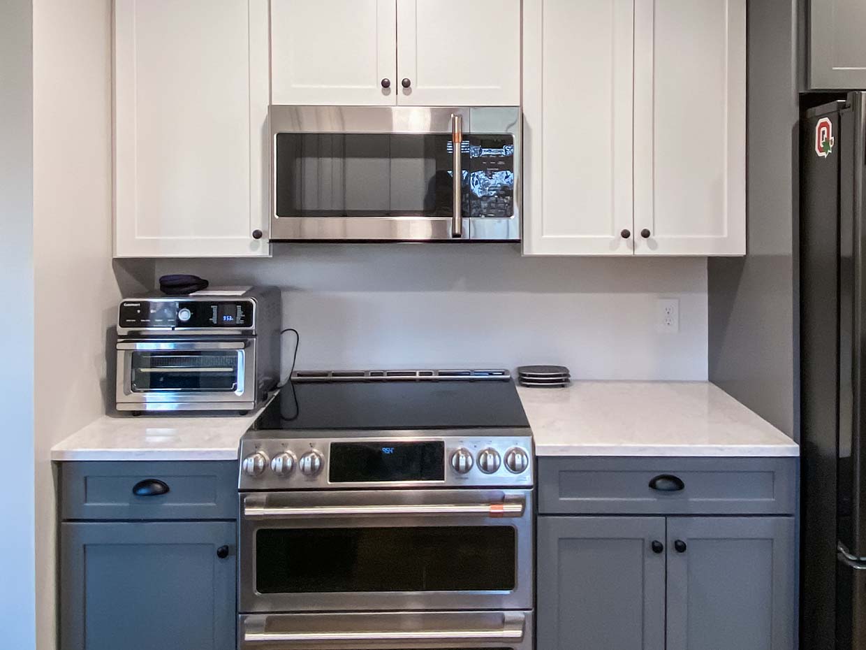 Modern kitchen with white upper cabinets, gray lower cabinets, stainless steel microwave, oven, toaster oven, and a black refrigerator. White countertops add contrast.