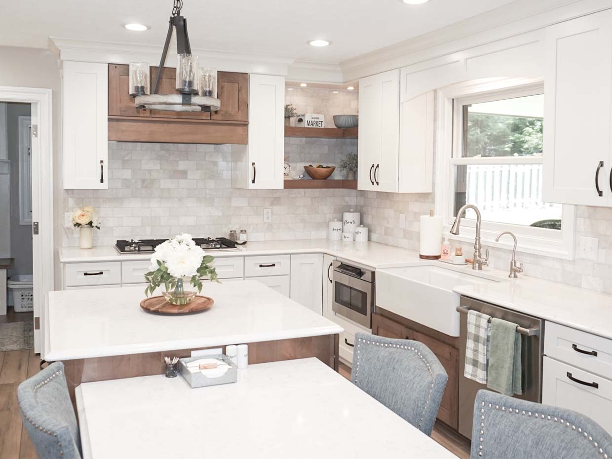 Modern kitchen with white cabinets, farmhouse sink, and a central island. Light wood accents, marble backsplash, and a chandelier complete the space.