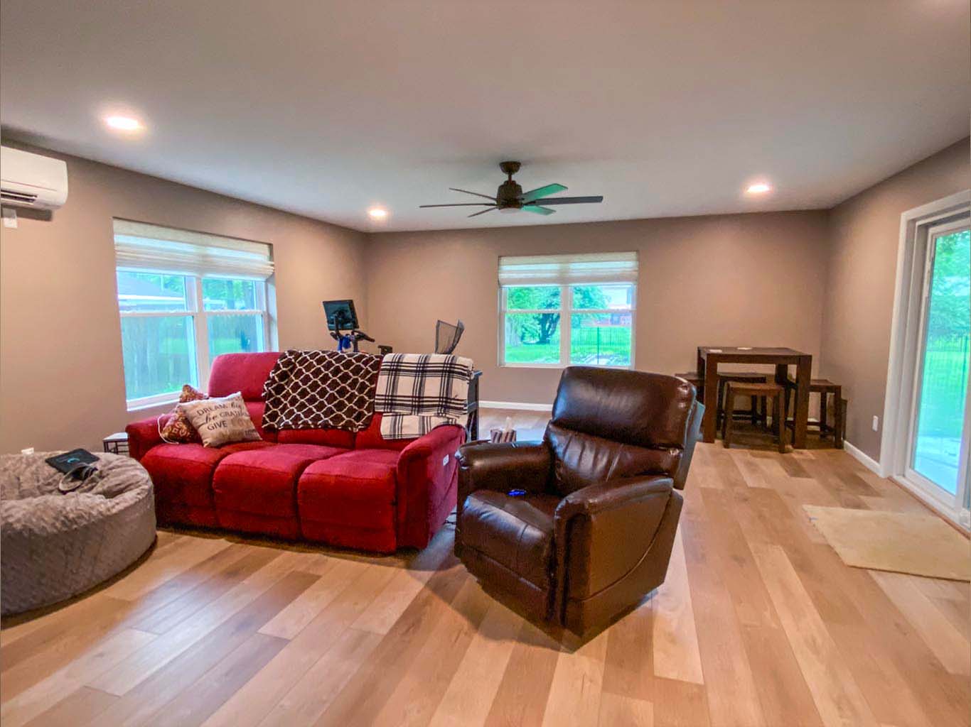 A living room with a red sofa, brown recliner, small dining table, and large windows. Ceiling fan and wood flooring are visible.